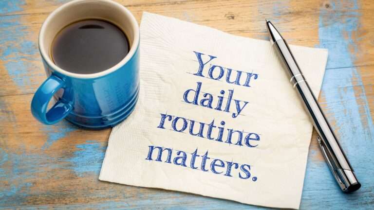 A wooden desk with a mug of coffee and pen resting on a note that reads "Your daily routine matters."