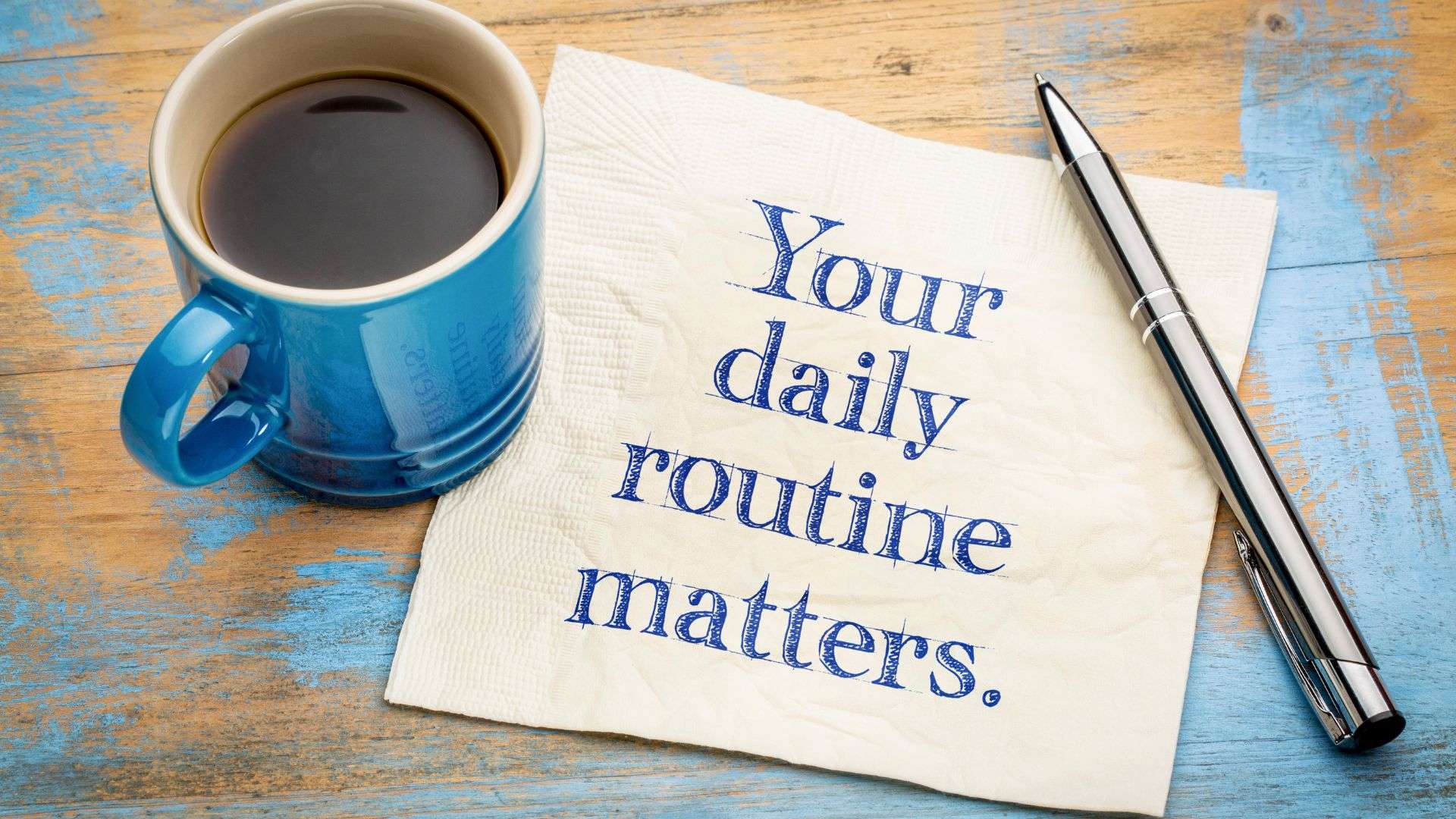 A wooden desk with a mug of coffee and pen resting on a note that reads "Your daily routine matters."