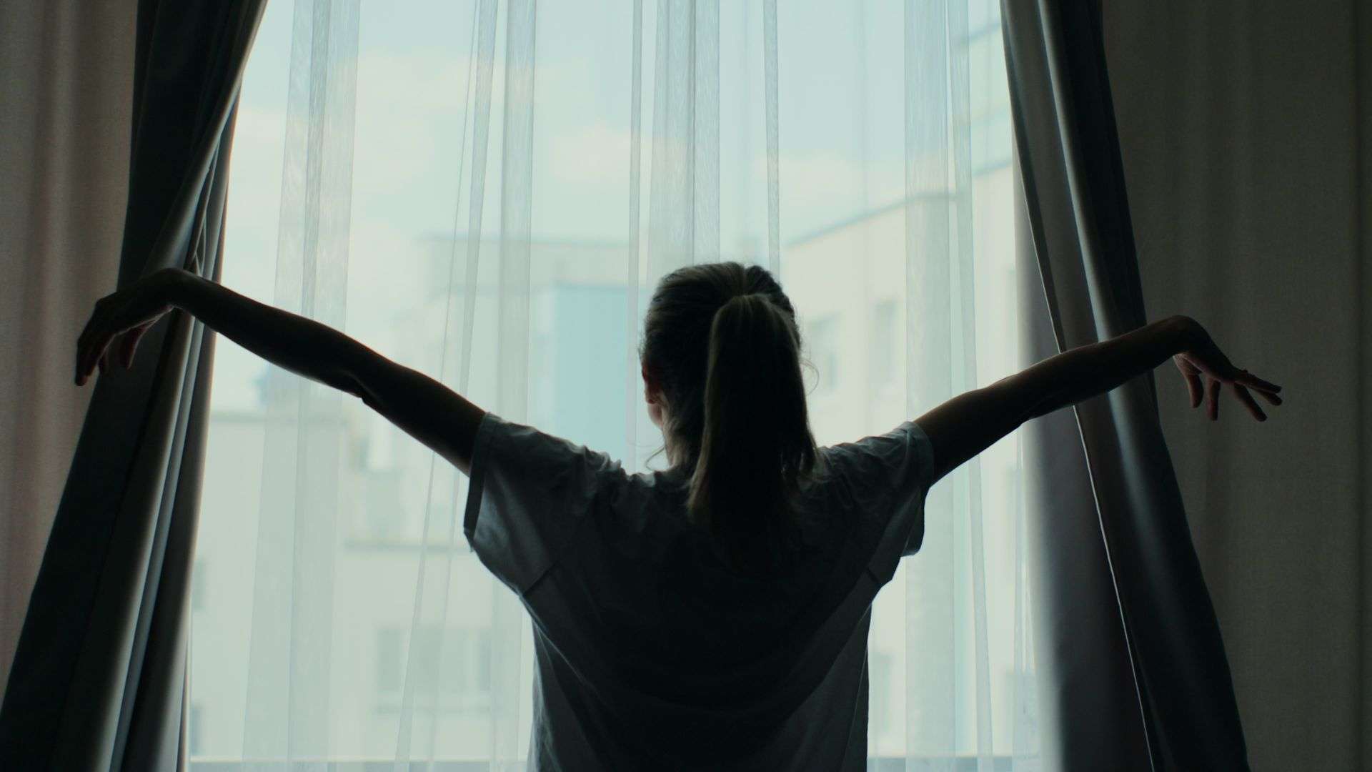 A woman standing at her window pulling the curtains back to see the sunrise.
