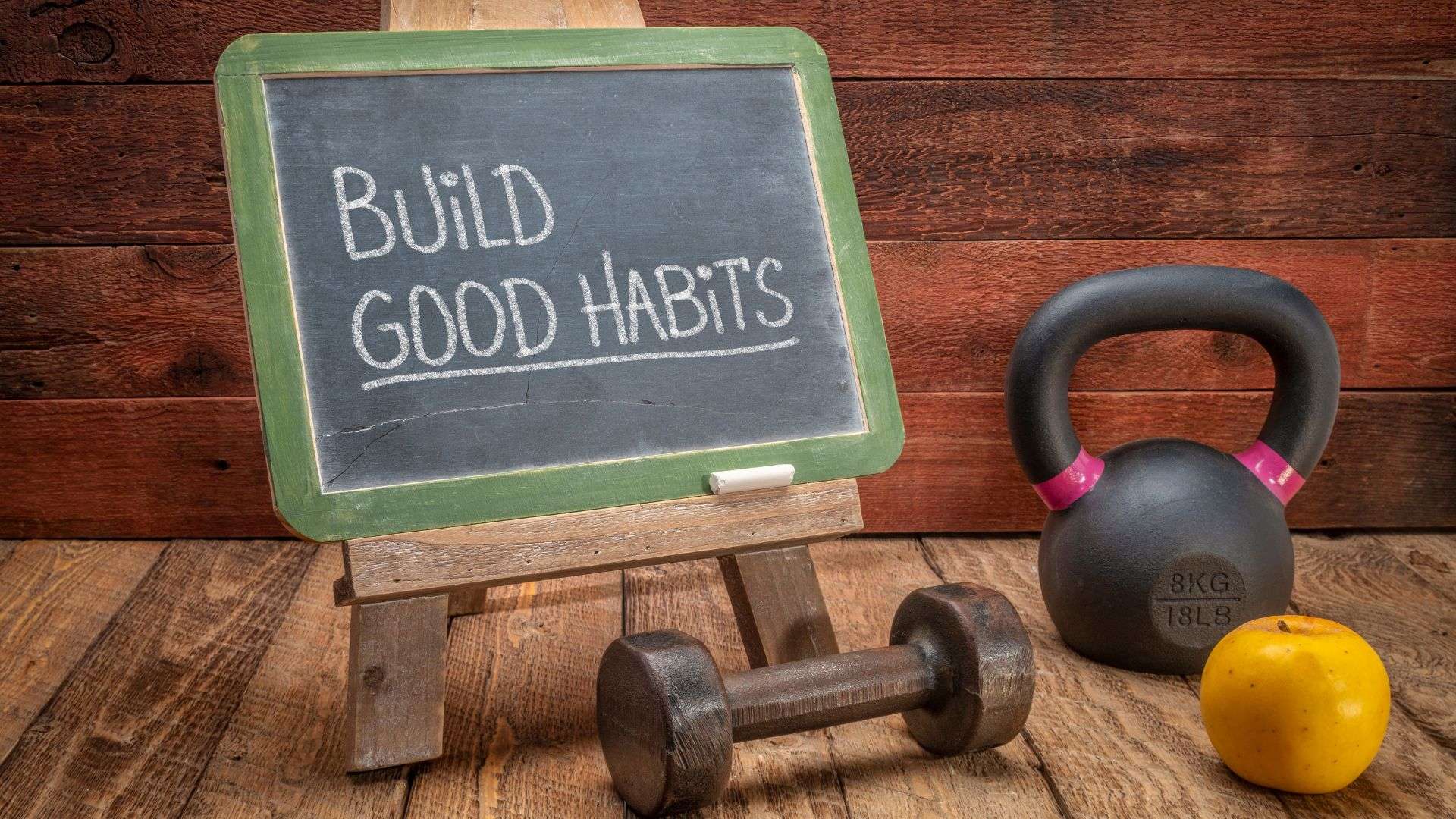 Chalkboard with the words "Build Good Habits" surrounded by a dumbbell, a kettle bell, and an apple.
